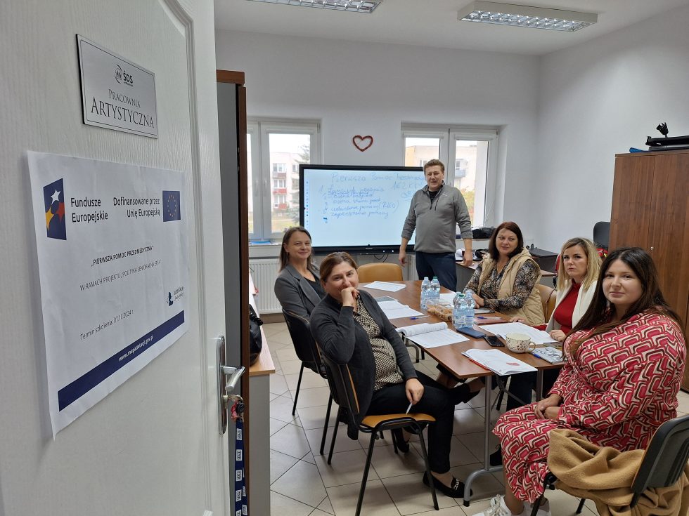 Zdjęcie przedstawia sześć osób w sali konferencyjnej. Pięć osób siedzi przy prostokątnym stole, jedna osoba stoi obok tablicy interaktywnej. Na stole znajdują się dokumenty, długopisy, plastikowe butelki z wodą oraz pudełko chusteczek higienicznych. W tle widoczne są dwa okna, przez które widać budynki znajdujące się na zewnątrz. Tablica interaktywna zawiera zapisane notatki. Na drzwiach do sali umieszczony jest plakat informacyjny dotyczący projektu finansowanego przez Fundusze Europejskie i Unię Europejską oraz tabliczka z napisem „Pracownia Artystyczna”.