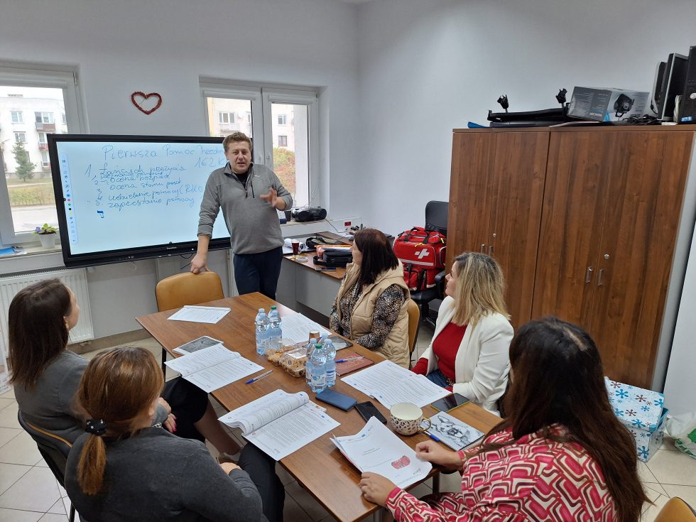 Zdjęcie przedstawia sześć osób w jasnym pomieszczeniu, siedzących przy prostokątnym stole. Na stole znajdują się dokumenty, notatki, długopisy oraz plastikowe butelki z wodą. Jedna osoba stoi obok tablicy interaktywnej, na której widoczny jest napis „Pierwsza Pomoc TEO”. Osoba stojąca wykonuje gest ręką w kierunku tablicy. W tle widoczne są dwa okna z widokiem na zewnętrzne budynki oraz drewniana szafa po prawej stronie. Na szafie umieszczone są różne przedmioty, w tym sprzęt elektroniczny i pudełka.