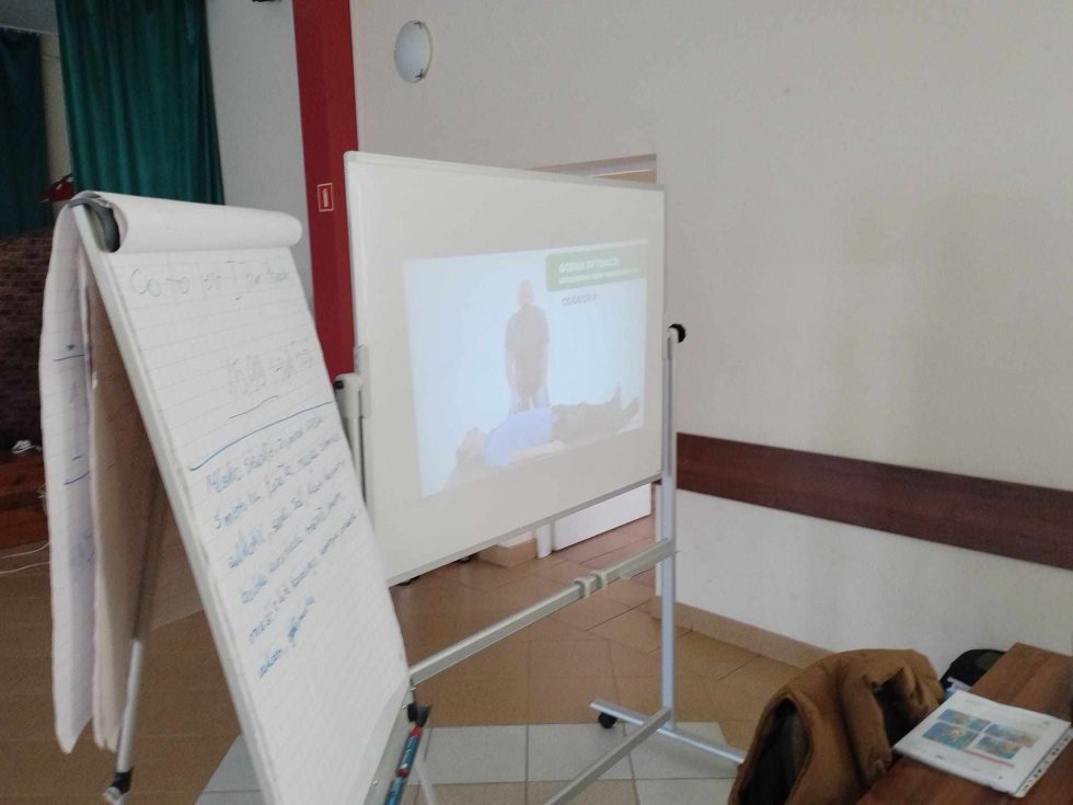 Zdjęcie przedstawia wnętrze sali szkoleniowej. Po lewej stronie znajduje się tablica typu flipchart z zapisanym tekstem w języku angielskim, zawierającym instrukcję „Go to next page then” oraz definicję stwardnienia rozsianego. W centralnej części zdjęcia widoczny jest ekran projekcyjny, na którym wyświetlony jest obraz przedstawiający osobę leżącą na podłodze oraz drugą osobę stojącą obok niej. Na ekranie znajduje się napis w języku polskim: „POZYCJA BEZPIECZNA”. Po prawej stronie zdjęcia widoczny jest stół, na którym leży brązowa kurtka oraz otwarty zeszyt lub książka.