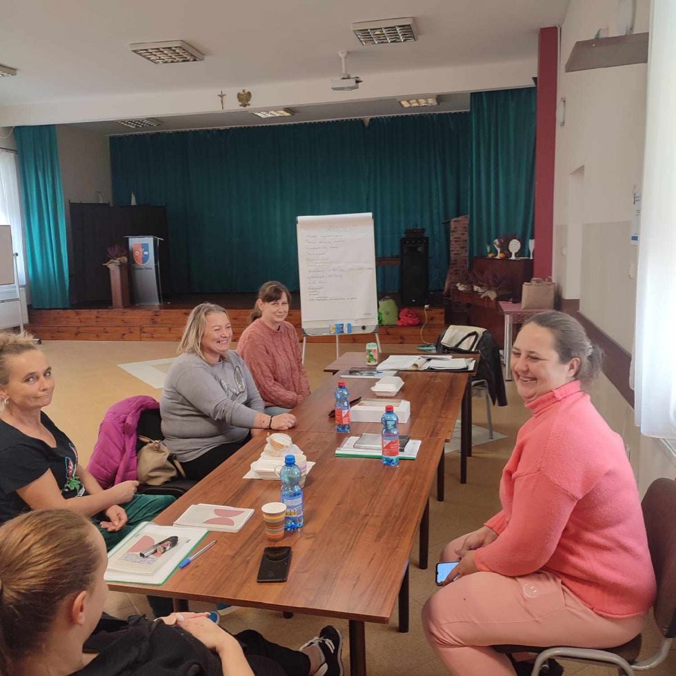 Zdjęcie przedstawia sześć osób siedzących przy prostokątnym, drewnianym stole w jasnym pomieszczeniu. Na stole znajdują się plastikowe butelki z wodą, kubki, notatniki oraz długopisy. W tle widoczna jest scena z zieloną zasłoną oraz tablica typu flipchart z zapisanymi notatkami.