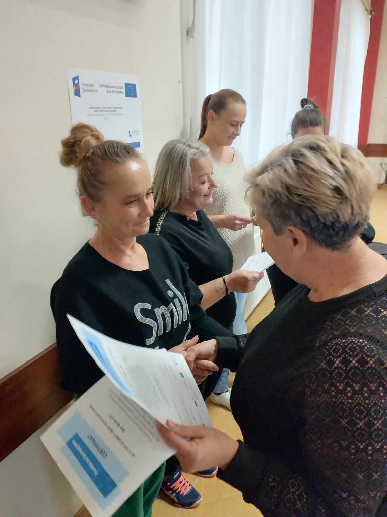 Zdjęcie przedstawia pięć osób stojących w rzędzie w jasnym pomieszczeniu. Wszystkie osoby trzymają dokumenty. Pierwsza osoba od lewej strony ubrana jest w czarną bluzę z napisem „Smile” i trzyma certyfikat. Druga osoba ma krótkie, siwe włosy i również trzyma dokument. Trzecia osoba ma związane włosy i jasny sweter. Czwarta osoba ubrana jest w ciemne ubranie i ma spięte włosy. Piąta osoba, znajdująca się najbliżej aparatu, jest zwrócona tyłem do obiektywu i przekazuje dokument pierwszej osobie od lewej. Na ścianie po lewej stronie znajduje się tablica informacyjna z logotypami, w tym logo Unii Europejskiej oraz napisem „Fundusze Europejskie”.