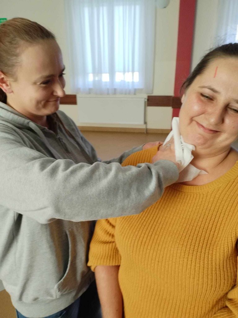 Zdjęcie przedstawia dwie osoby w pomieszczeniu. Osoba po lewej stronie ubrana jest w szarą bluzę z kapturem i trzyma biały materiał lub chusteczkę, którą przykłada do szyi osoby stojącej po prawej stronie. Osoba po prawej stronie ubrana jest w żółty sweter i stoi bokiem do aparatu. W tle widoczne są zasłonięte białe firanki na oknach oraz fragment czerwonego filaru.