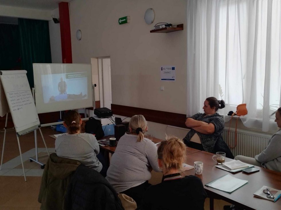 Zdjęcie przedstawia pięć osób siedzących przy prostokątnym stole w sali. Osoby są zwrócone w stronę ekranu projekcyjnego, na którym wyświetlany jest film instruktażowy pokazujący osobę wykonującą ćwiczenia na macie. Obok ekranu znajduje się tablica typu flipchart z zapisanymi notatkami. Na stole rozmieszczone są kubki z napojami, notesy oraz długopisy. W tle widoczne są białe zasłony na oknach, półka z urządzeniami elektronicznymi oraz plakat informacyjny zawieszony na ścianie obok okna.