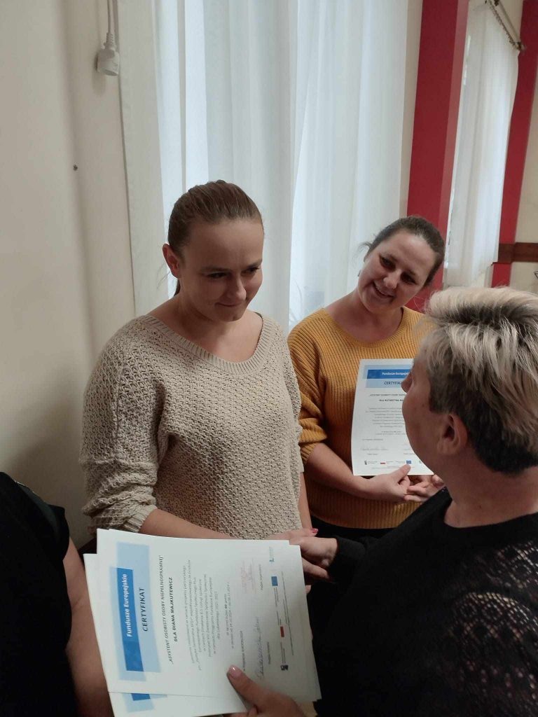 Zdjęcie przedstawia cztery osoby stojące w pomieszczeniu. Trzy osoby trzymają certyfikaty, a jedna osoba wręcza dokumenty pozostałym. Certyfikaty zawierają niebieskie i białe elementy graficzne oraz tekst, który jest częściowo widoczny. W tle znajdują się białe zasłony na oknach oraz fragment czerwonej ściany.