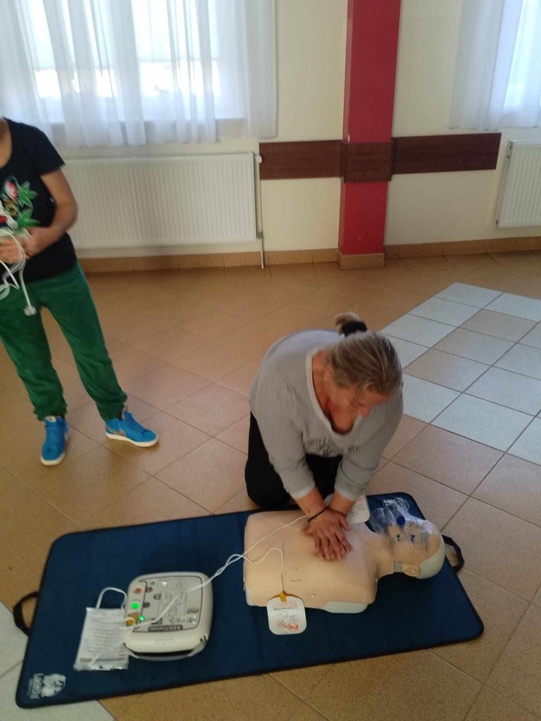 Zdjęcie przedstawia osobę wykonującą resuscytację krążeniowo-oddechową (RKO) na manekinie treningowym. Osoba klęczy na podłodze i umieszcza dłonie na klatce piersiowej manekina, wykonując uciski. Manekin leży na niebieskiej macie treningowej. Obok manekina znajduje się automatyczny defibrylator zewnętrzny (AED), podłączony do manekina za pomocą elektrod. W tle widoczna jest druga osoba stojąca obok, trzymająca przewody AED. Pomieszczenie ma jasne ściany, duże okna z białymi zasłonami oraz grzejniki umieszczone pod oknami.