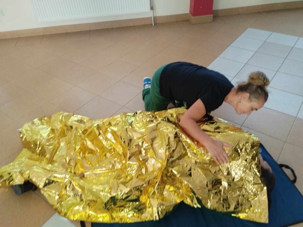 Zdjęcie przedstawia osobę klęczącą na podłodze w pomieszczeniu, która przykrywa drugą osobę folią ratunkową w kolorze złotym. Osoba leżąca znajduje się na niebieskim materacu, a folia zakrywa całe jej ciało. Podłoga wykonana jest z jasnobrązowych płytek.