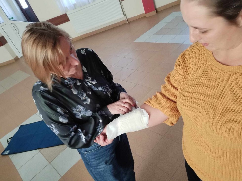 Zdjęcie przedstawia dwie osoby znajdujące się w pomieszczeniu z jasnobrązową podłogą i białymi ścianami. Osoba po lewej stronie, ubrana w ciemną koszulę z kwiatowym wzorem oraz niebieskie spodnie, owija bandażem przedramię osoby stojącej po prawej stronie. Osoba po prawej stronie ma na sobie żółty sweter i czarne spodnie. W tle widoczne są drzwi, okno z zasłonami oraz biały kaloryfer. Na podłodze znajduje się niebieska mata do ćwiczeń.