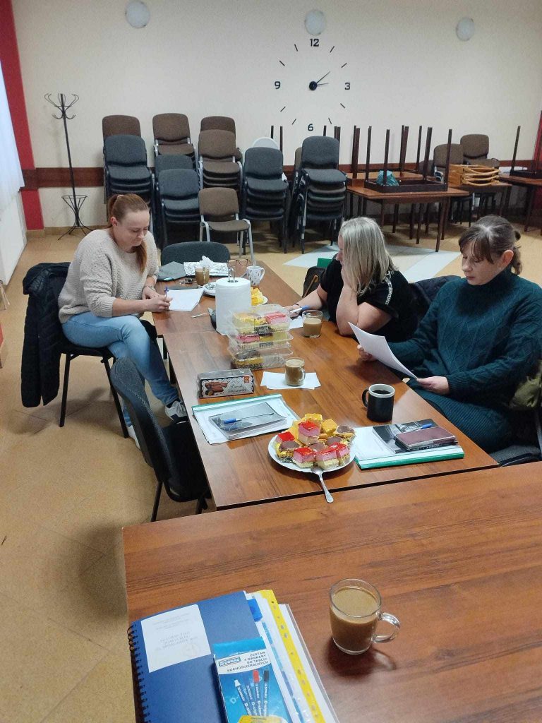 Zdjęcie przedstawia salę konferencyjną, w której trzy osoby siedzą przy długim, drewnianym stole. Na stole znajdują się filiżanki z napojami, talerz z ciastkami, dokumenty, notatki oraz inne drobne przedmioty. W tle widoczne są stosy plastikowych krzeseł ustawionych jedno na drugim, zegar ścienny wskazujący godzinę 9:05 oraz stojak na ubrania umieszczony w lewym rogu pomieszczenia.