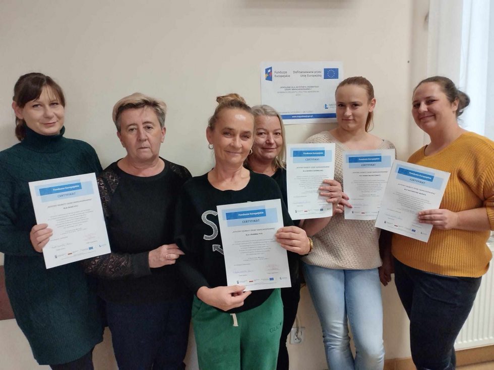 Zdjęcie przedstawia sześć osób stojących w rzędzie na tle jasnej ściany. Każda z osób trzyma certyfikat z niebieskim nagłówkiem i tekstem poniżej. Dokumenty zawierają słowo „CERTYFIKAT” oraz logotypy umieszczone w dolnej części. Na ścianie za osobami znajduje się plakat informacyjny z logotypami, w tym logo Unii Europejskiej, oraz tekstem dotyczącym projektu finansowanego ze środków Unii Europejskiej.