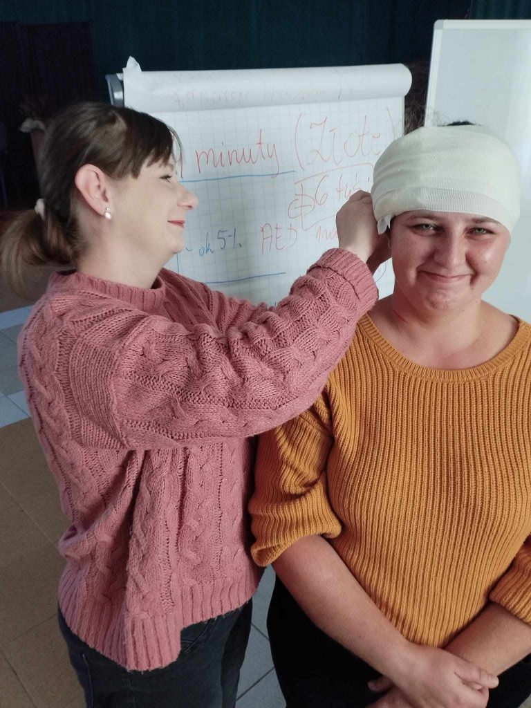 Zdjęcie przedstawia dwie osoby znajdujące się w pomieszczeniu. Osoba po lewej stronie, ubrana w różowy sweter, zakłada bandaż na głowę osoby po prawej stronie, która ma na sobie musztardowy sweter. W tle znajduje się biała tablica typu flipchart ze stojakiem, na której zapisane są notatki w języku polskim.