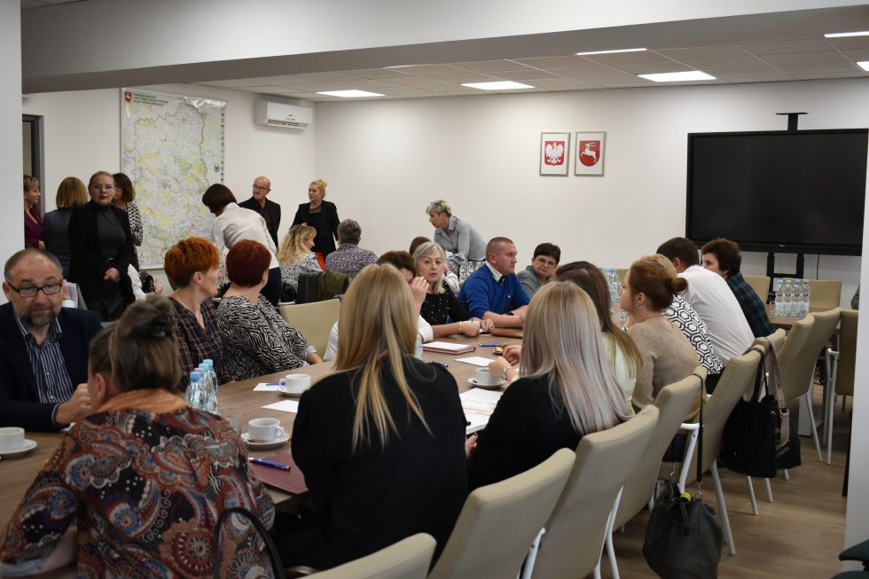 Zdjęcie przedstawia salę konferencyjną z dużym stołem, przy którym siedzi grupa osób. Na stole znajdują się filiżanki z kawą, butelki z wodą oraz dokumenty. W tle widoczna jest mapa zawieszona na ścianie oraz dwie ramki z symbolami państwowymi Polski. Po prawej stronie znajduje się duży ekran telewizyjny zamontowany na stojaku. W pomieszczeniu obecne są również osoby stojące i rozmawiające. Sala jest dobrze oświetlona, a na suficie zamontowane są panele świetlne.