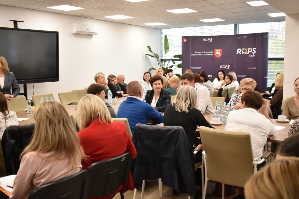 Zdjęcie przedstawia salę konferencyjną z grupą osób siedzących przy dużym stole. Na stole znajdują się butelki z wodą, filiżanki oraz dokumenty. W tle widoczne są dwa banery z logotypami i napisami „ROPS Lublin” oraz „Województwo Lubelskie”. Po prawej stronie znajdują się duże okna, przez które wpada naturalne światło. Przy oknach ustawione są rośliny doniczkowe. Na lewej ścianie znajduje się duży ekran telewizyjny lub monitor. Uczestnicy spotkania są różnie ubrani i zajmują się rozmowami lub notowaniem.