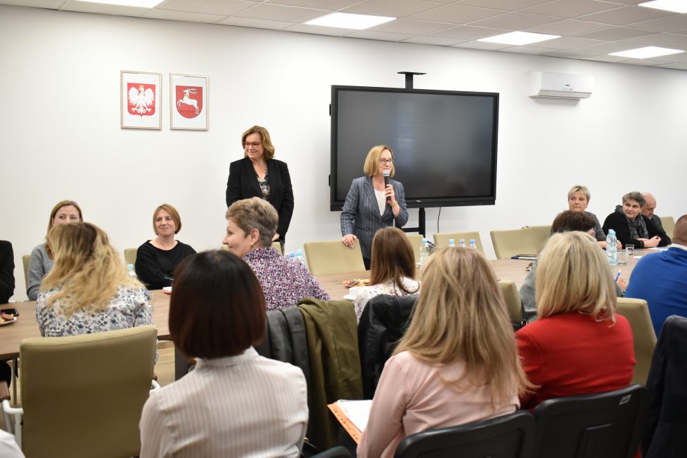 Zdjęcie przedstawia grupę osób siedzących przy dużym, prostokątnym stole konferencyjnym w sali spotkań. Na pierwszym planie widoczne są osoby siedzące tyłem do aparatu. W dalszej części zdjęcia znajdują się osoby siedzące przodem do obiektywu. Na ścianie w tle, centralnie, zamontowany jest duży ekran telewizyjny. Po prawej stronie ekranu stoją dwie osoby: jedna trzyma mikrofon i przemawia do zgromadzonych, druga również zwraca się do uczestników spotkania.