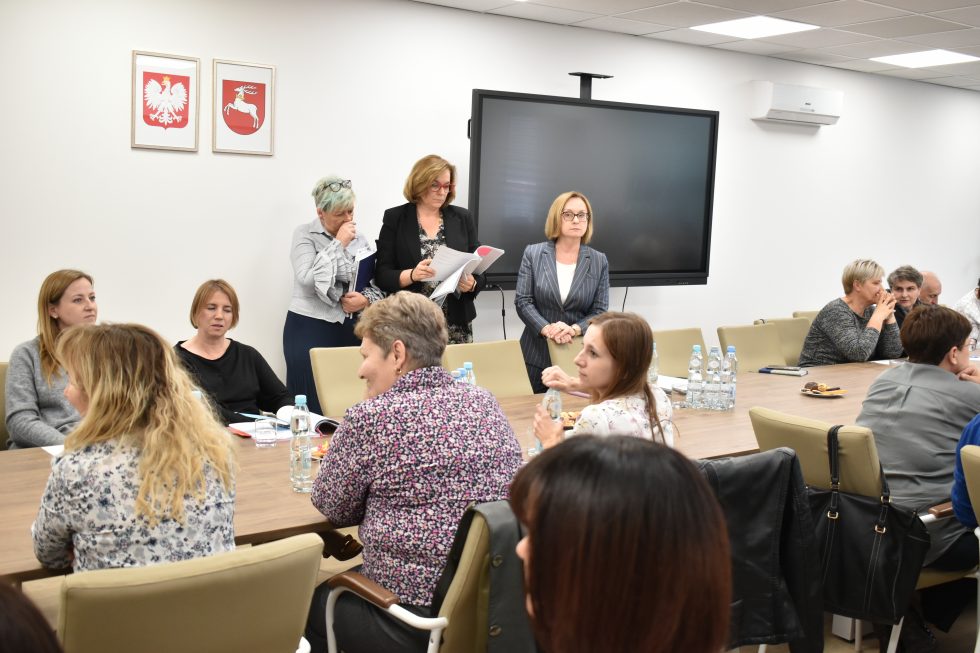 Grupa osób siedzi przy dużym, prostokątnym, drewnianym stole w sali konferencyjnej. Na pierwszym planie widoczne są osoby siedzące tyłem i bokiem do obiektywu. W dalszej części zdjęcia znajdują się osoby zwrócone przodem do aparatu. Po prawej stronie znajduje się duży ekran telewizyjny lub monitor zawieszony na ścianie. Na powierzchni stołu rozmieszczone są plastikowe butelki z wodą mineralną, dokumenty, notatniki oraz inne materiały papierowe.