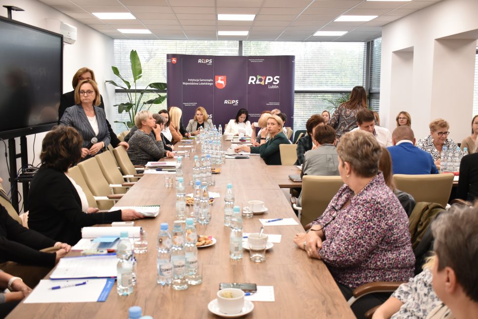 Zdjęcie przedstawia salę konferencyjną z grupą osób siedzących przy dużym stole. Na stole znajdują się butelki z wodą, filiżanki oraz dokumenty. W tle widoczne są dwa banery z logotypami i napisami „ROPS Lublin” oraz „Województwo Lubelskie”. Po prawej stronie znajdują się duże okna, przez które wpada naturalne światło. Przy oknach ustawione są rośliny doniczkowe. Na lewej ścianie znajduje się duży ekran telewizyjny lub monitor. Uczestnicy spotkania są różnie ubrani i zajmują się rozmowami lub notowaniem.