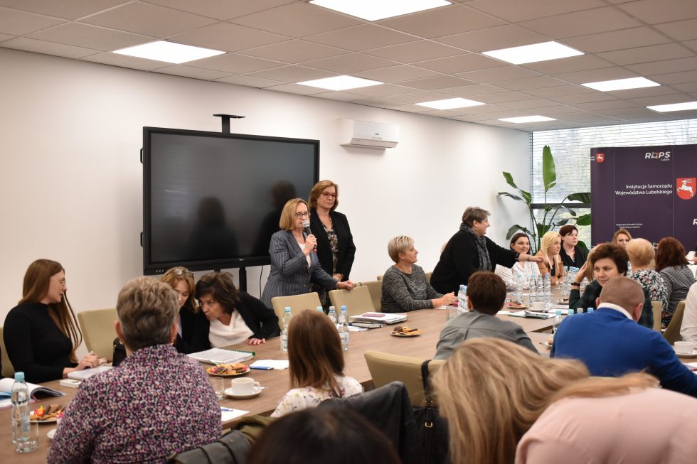 Zdjęcie przedstawia salę konferencyjną z grupą osób siedzących przy dużym stole. Na stole znajdują się butelki z wodą, filiżanki oraz dokumenty. W tle widoczne są dwa banery z logotypami i napisami „ROPS Lublin” oraz „Województwo Lubelskie”. Po prawej stronie znajdują się duże okna, przez które wpada naturalne światło. Przy oknach ustawione są rośliny doniczkowe. Na lewej ścianie znajduje się duży ekran telewizyjny lub monitor. Uczestnicy spotkania są różnie ubrani i zajmują się rozmowami lub notowaniem.