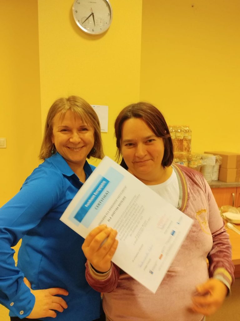 Zdjęcie przedstawia dwie osoby stojące w pomieszczeniu o żółtych ścianach. Osoba po lewej stronie ubrana jest w niebieską koszulę. Osoba po prawej stronie trzyma certyfikat w dłoni. W tle znajduje się zegar ścienny oraz stół, na którym ustawione są różne przedmioty, w tym słoiki i pudełka.