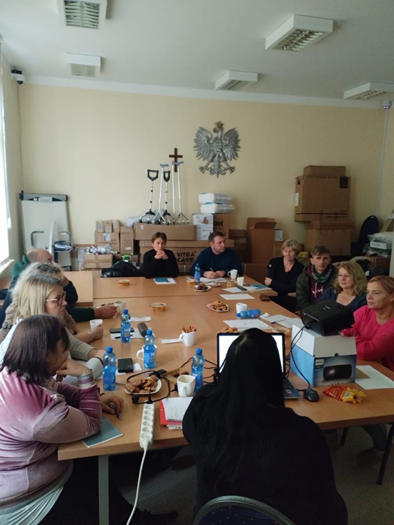 Zdjęcie przedstawia grupę osób siedzących przy dużym, prostokątnym stole w sali konferencyjnej. Na stole znajdują się plastikowe butelki z wodą, filiżanki, talerze z przekąskami, dokumenty, notatniki oraz długopisy. Jedna osoba korzysta z laptopa. W tle widoczne są kartony, kule ortopedyczne, krzyż oraz godło Polski zawieszone na ścianie.