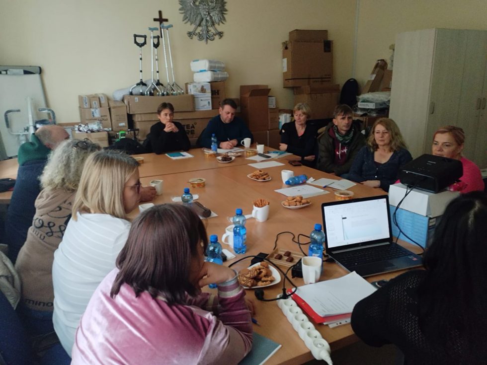 Zdjęcie przedstawia grupę osób siedzących wokół dużego, prostokątnego stołu w sali konferencyjnej. Na stole znajdują się plastikowe butelki z wodą, filiżanki z napojami, talerze z przekąskami, notatniki oraz długopisy. Na jednym końcu stołu ustawiony jest laptop podłączony do projektora, który wyświetla slajd na ekranie. W tle widoczne są kartony, kule ortopedyczne, szafa oraz dekoracje ścienne.