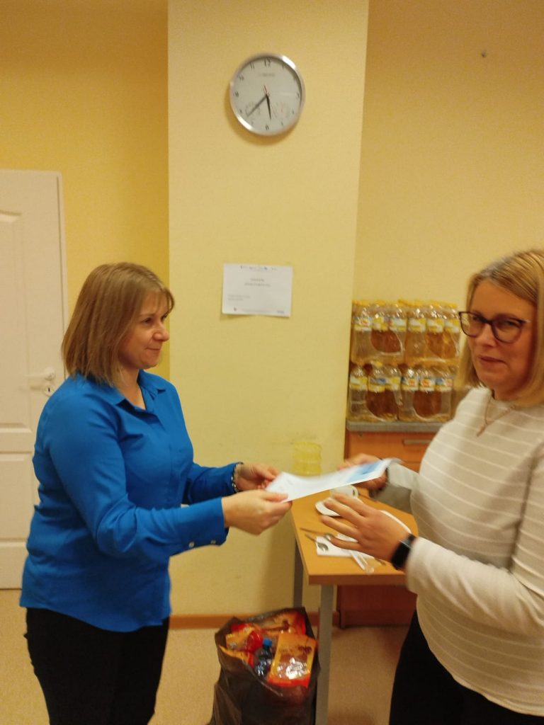 Zdjęcie przedstawia dwie osoby stojące w pomieszczeniu. Osoba po lewej stronie ubrana jest w niebieską koszulę i trzyma dokument, który przekazuje osobie po prawej stronie. Osoba po prawej stronie ma na sobie jasny sweter i okulary, wyciąga ręce w kierunku dokumentu.