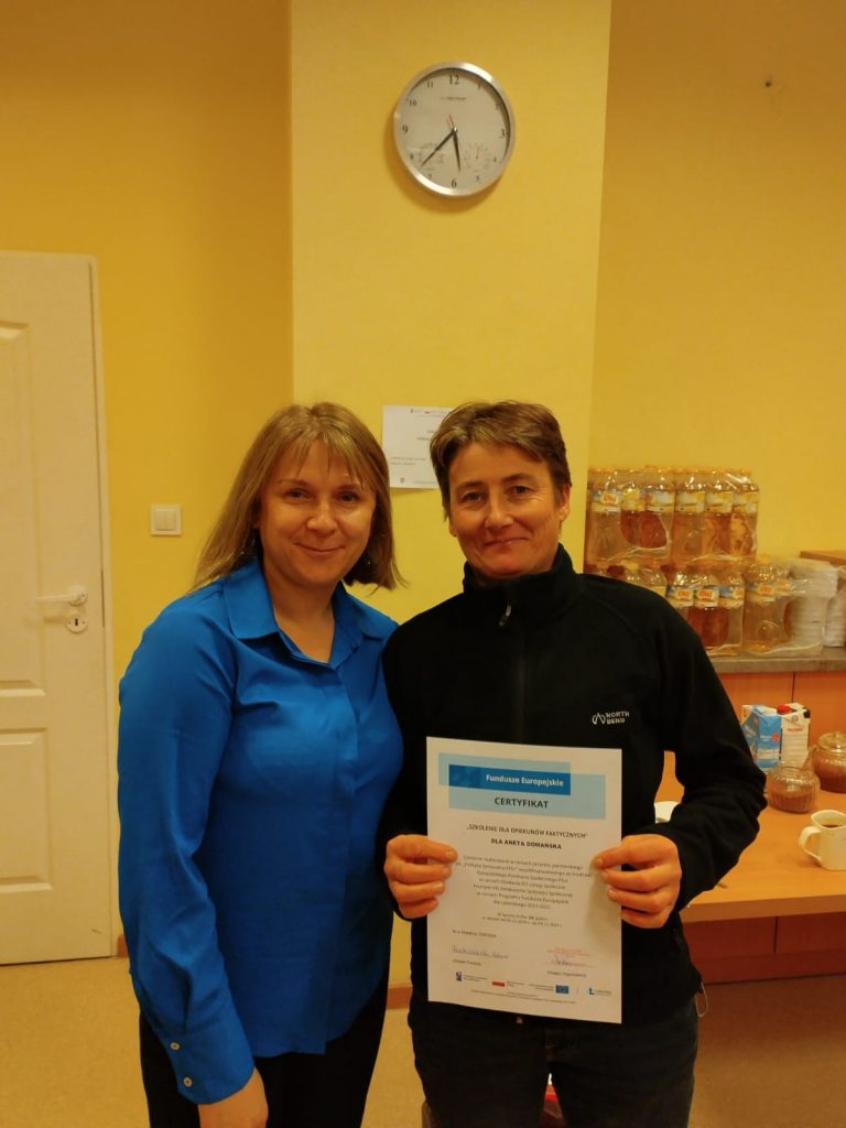 Zdjęcie przedstawia dwie osoby stojące obok siebie w pomieszczeniu. Osoba po lewej stronie ubrana jest w niebieską koszulę. Osoba po prawej stronie trzyma certyfikat oburącz. Dokument zawiera nagłówek „CERTYFIKAT” oraz informacje o przyznaniu certyfikatu przez „Fundusze Europejskie”.