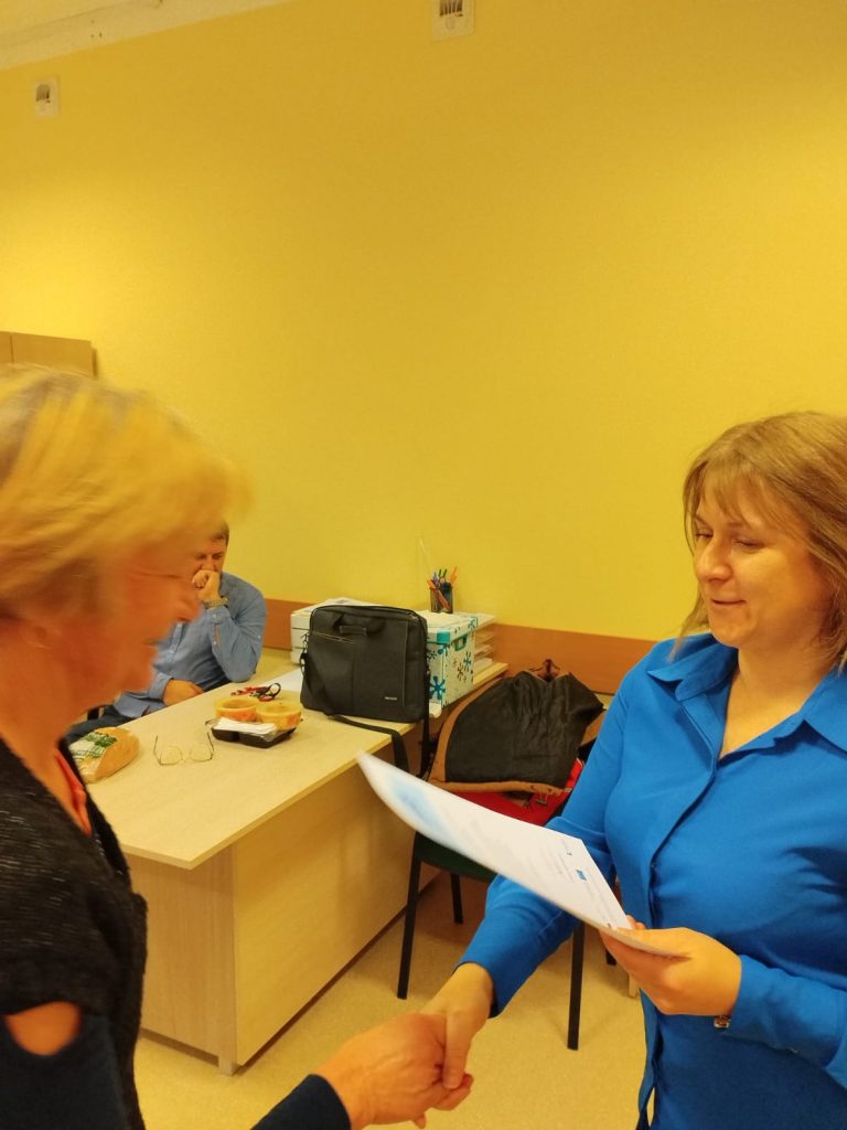 Zdjęcie przedstawia dwie osoby znajdujące się w pomieszczeniu o żółtych ścianach. Osoba po prawej stronie, ubrana w niebieską koszulę, trzyma dokument i przekazuje go osobie po lewej stronie, która jest częściowo widoczna. W tle znajduje się biurko, na którym umieszczone są różne przedmioty, w tym torba, pudełka z chusteczkami oraz przybory do pisania. Za biurkiem, na krześle, siedzi trzecia osoba również ubrana w niebieską koszulę.