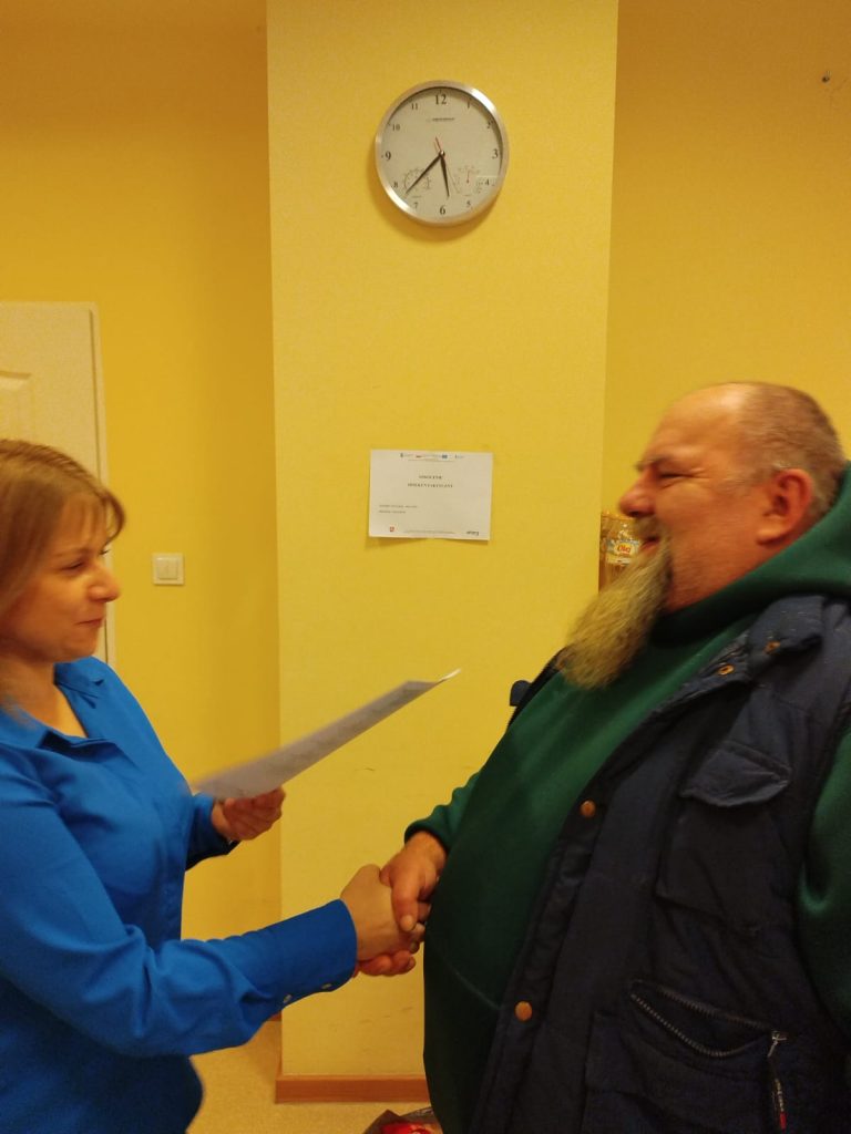 Zdjęcie przedstawia dwie osoby stojące naprzeciwko siebie w pomieszczeniu o żółtych ścianach. Osoba po lewej stronie ubrana jest w niebieską koszulę, trzyma dokument i podaje rękę osobie po prawej stronie. Osoba po prawej stronie ma na sobie zieloną bluzę z kapturem oraz granatową kamizelkę.