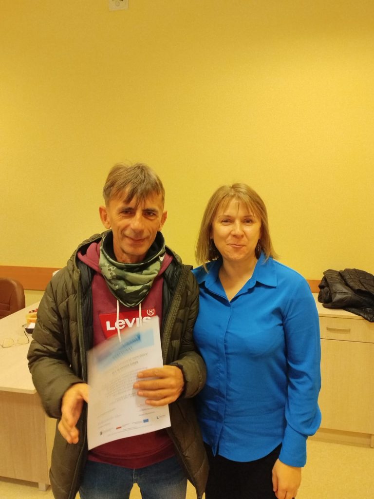 Zdjęcie przedstawia dwie osoby stojące obok siebie w pomieszczeniu. Osoba po lewej stronie trzyma dokument w rękach. Osoba ta ubrana jest w ciemną kurtkę, bordową bluzę z kapturem z widocznym logo marki Levi’s oraz zielony szalik. Osoba po prawej stronie ma na sobie niebieską koszulę i czarne spodnie. W tle widoczna jest jasna ściana, stół z kilkoma przedmiotami oraz szafka złożona z kilku szuflad, na której leży ciemna kurtka.