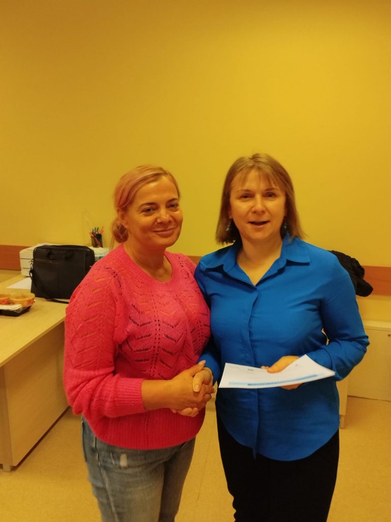 Zdjęcie przedstawia dwie osoby stojące w pomieszczeniu. Osoba po lewej stronie ubrana jest w różowy sweter i trzyma rękę osoby po prawej stronie. Osoba po prawej stronie ma na sobie niebieską koszulę i trzyma dokumenty. W tle widoczne jest biurko, na którym znajduje się czarna torba oraz przybory do pisania.