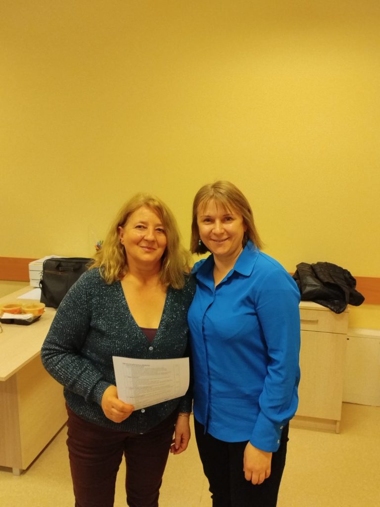 Zdjęcie przedstawia dwie osoby stojące obok siebie w pomieszczeniu. Osoba po lewej stronie trzyma kartkę papieru z tekstem. W tle widoczne są biurka z jasnym drewnianym wykończeniem. Na jednym z biurek znajduje się czarna torba oraz pudełka z jedzeniem.