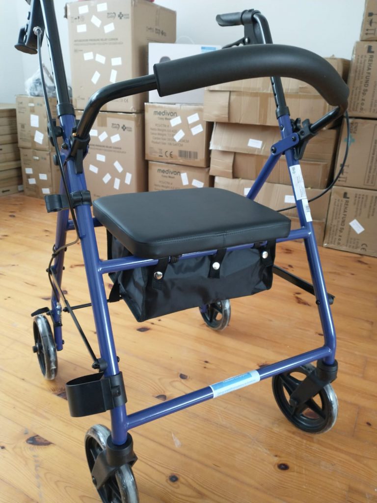 Niebieski chodzik czterokołowy z siedziskiem i torbą na zakupy, ustawiony na drewnianej podłodze w pomieszczeniu. W tle widoczne są kartony z etykietami „medivon”.