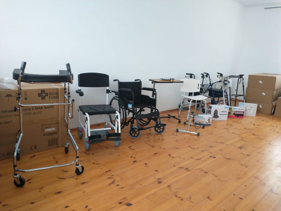 Pomieszczenie z drewnianą podłogą, w którym ustawiono urządzenia medyczne. Od lewej strony widoczny jest chodzik na kółkach, krzesło toaletowe, wózek inwalidzki, stolik przyłóżkowy, krzesło prysznicowe oraz trzy chodziki. W tle po prawej stronie znajdują się kartony i pudełka z produktami medycznymi.