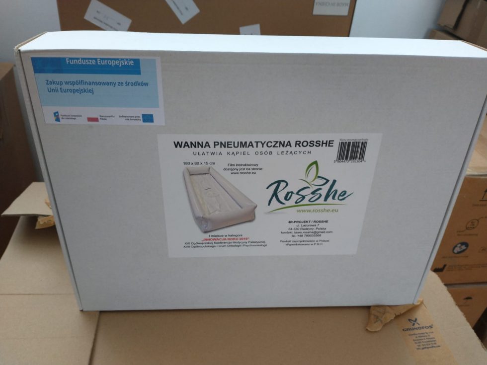 Na zdjęciu znajduje się kartonowe pudełko z nadrukiem przedstawiającym produkt – wannę pneumatyczną marki Rosshe. Na froncie pudełka widnieje duży napis „WANNA PNEUMATYCZNA ROSSHE” oraz logo producenta. W lewym górnym rogu pudełka umieszczono informację o współfinansowaniu zakupu ze środków Unii Europejskiej.
