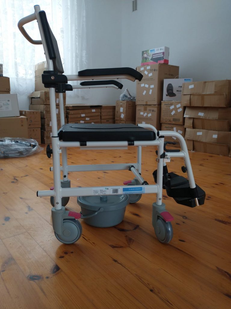 Krzesło toaletowe na kółkach, ustawione na drewnianej podłodze w pomieszczeniu. Konstrukcja krzesła wykonana z metalu, z czarnym siedziskiem, oparciem i bocznymi uchwytami. Pod siedziskiem znajduje się plastikowy pojemnik. W tle widoczne są stosy kartonów i pudełek oraz zasłonięte okno z firanką po lewej stronie.