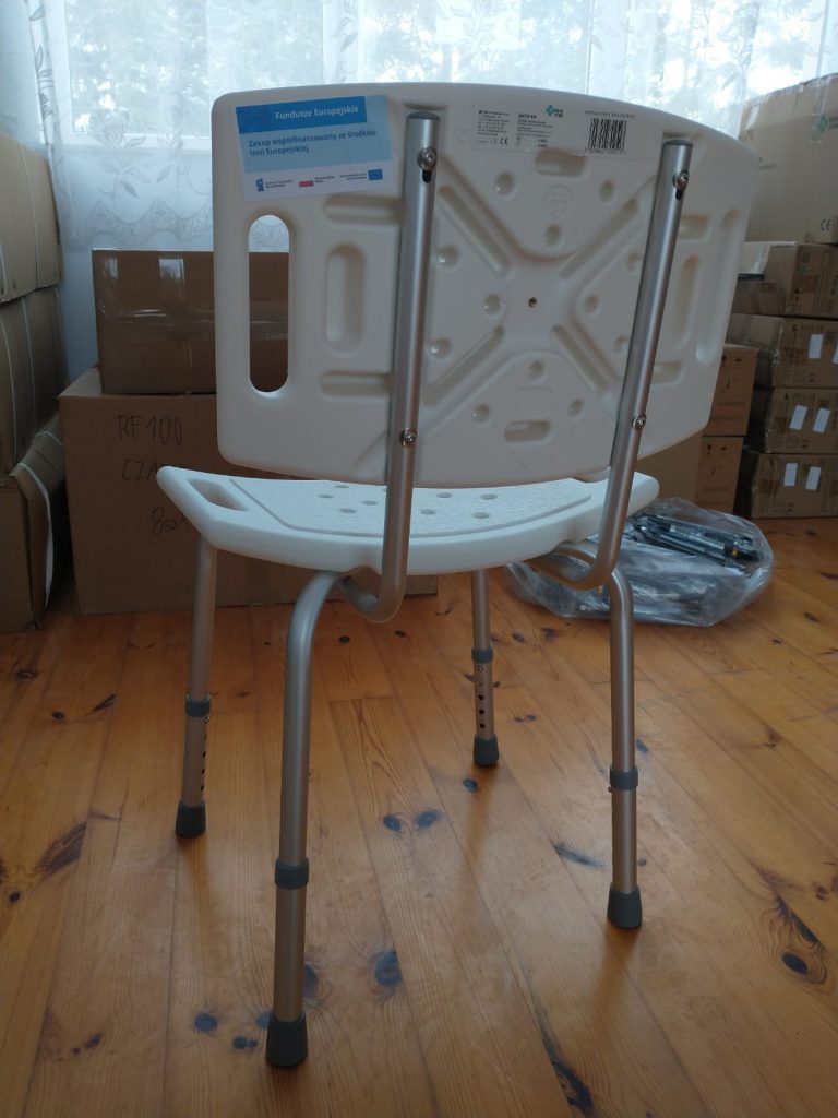 Plastikowe krzesło z metalowymi nogami, ustawione na drewnianej podłodze. Krzesło posiada otwory w oparciu i siedzisku oraz regulowane nóżki zakończone gumowymi nakładkami. Na oparciu znajduje się niebieska etykieta z napisem „Fundacja Kresy-Syberia” oraz informacją o projekcie współfinansowanym ze środków Unii Europejskiej. W tle widoczne są kartony, pudełka oraz zasłonięte firanką okno.