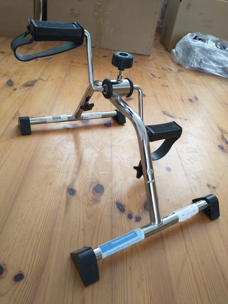Metalowy rowerek treningowy do ćwiczeń kończyn dolnych i górnych, ustawiony na drewnianej podłodze. Wyposażony w dwa pedały z czarnymi paskami na stopy oraz uchwyt z czarnym paskiem na ręce. Na centralnej części urządzenia znajduje się pokrętło do regulacji oporu. W tle widoczne są kartony i plastikowa torba.