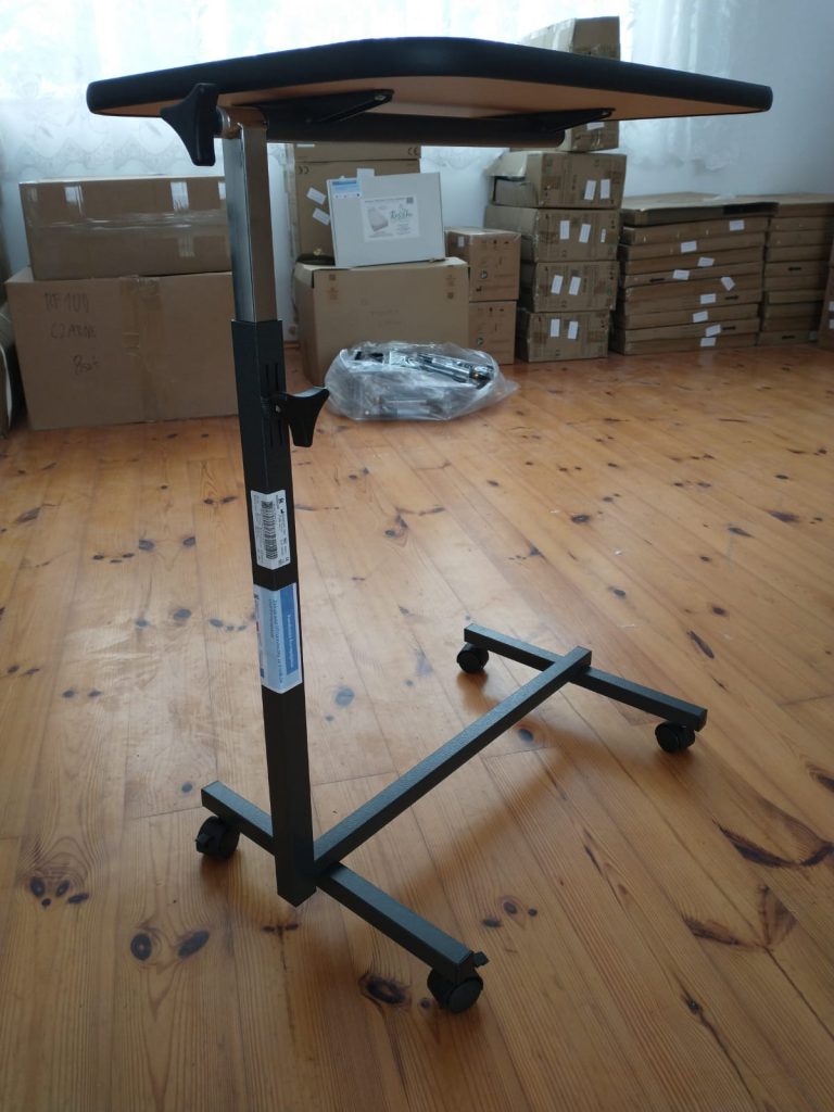 Mobilny stolik na kółkach z regulowaną wysokością, posiadający metalową ramę w kształcie litery „T” z czterema kółkami u podstawy. Blat stolika jest prostokątny, wykonany z drewna lub materiału drewnopodobnego, z lekko nachyloną powierzchnią.