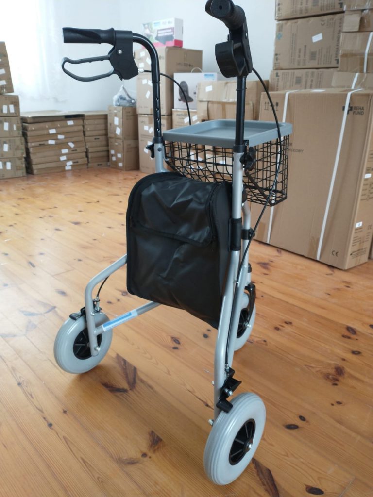 Chodzik na czterech kołach z ręcznymi hamulcami, wyposażony w koszyk na zakupy i torbę. Urządzenie ustawione na drewnianej podłodze w pomieszczeniu, w którym znajdują się stosy kartonów.