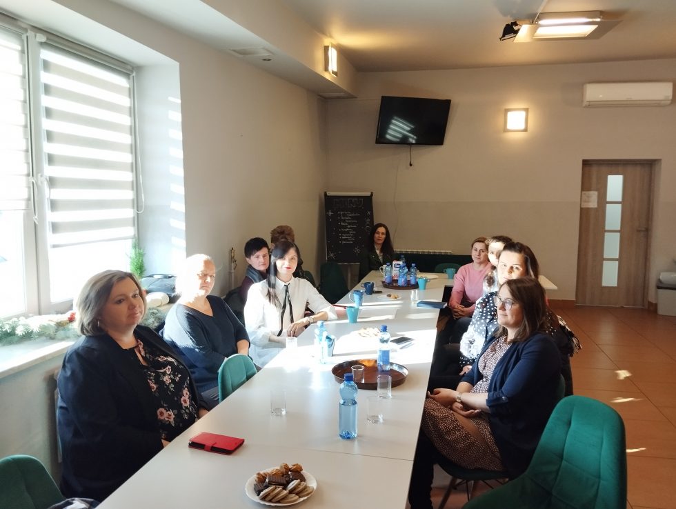 Zdjęcie przedstawia osoby, kobiety, siedzące przy stole w małej sali konferencyjnej.