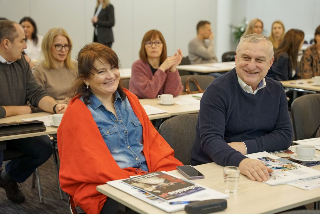 Uczestnicy siedzą przy stolikach. Przed nimi notesy, dokumenty i kubki.
