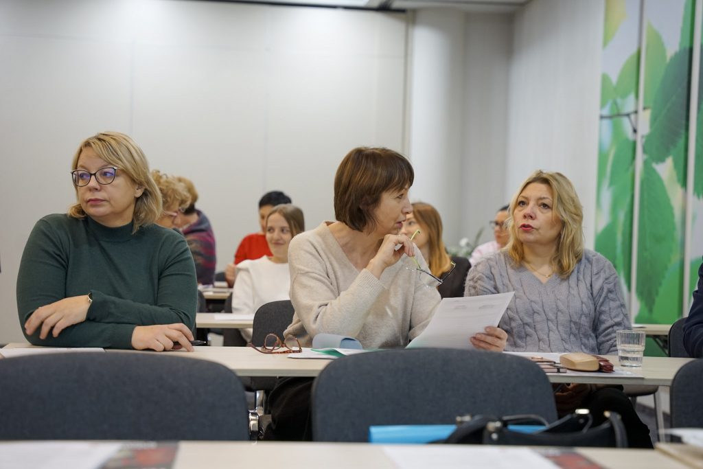 Kilka osób siedzi w sali, jedna trzyma kartkę, inne słuchają lub patrzą w bok.
