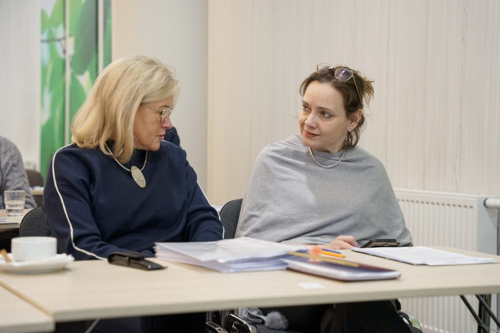 Dwie osoby rozmawiają przy stoliku. Na blacie filiżanki, zeszyty i materiały.