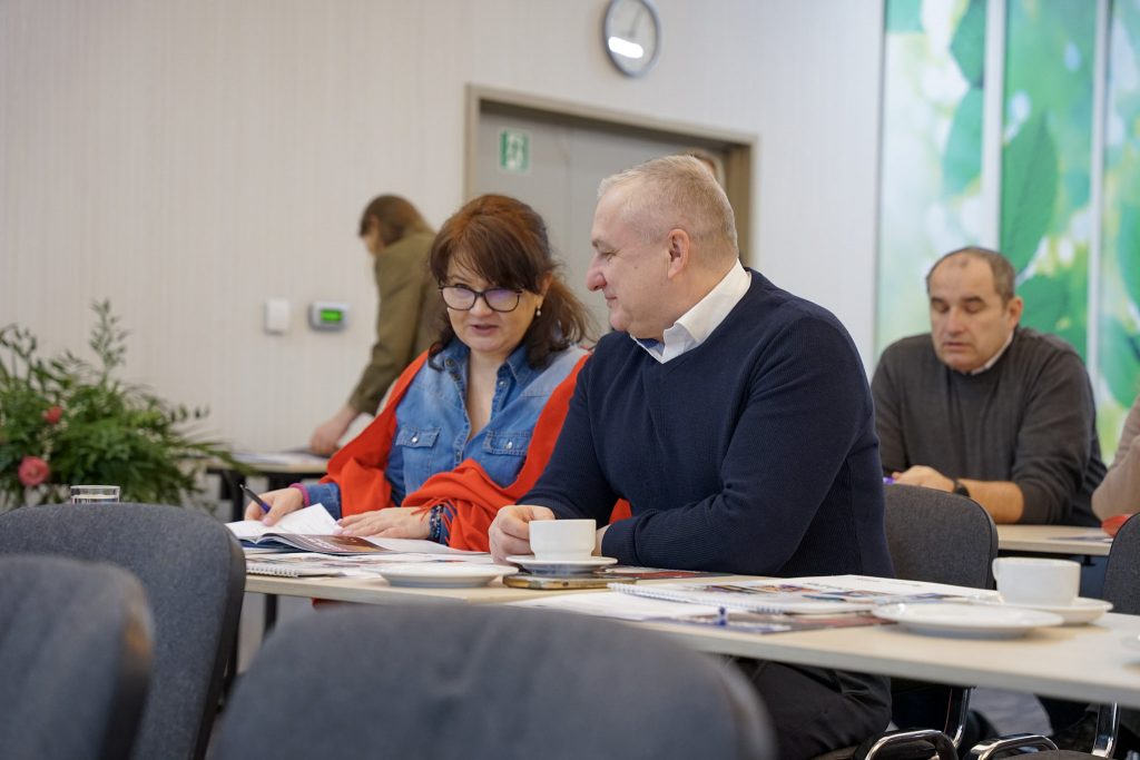 Kobieta gestykuluje podczas rozmowy z siedzącym obok mężczyzną. Na stole leżą dokumenty.
