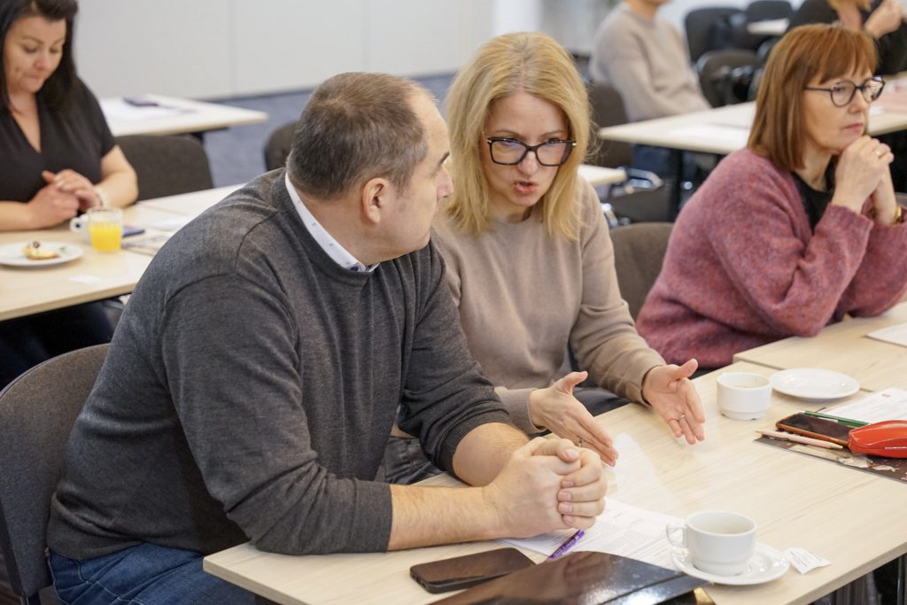 Kobieta i mężczyzna siedzą przy stoliku i rozmawiają. Na stole leżą dokumenty, długopis i filiżanka.