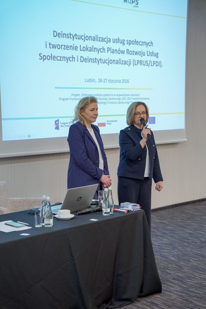 Dwie kobiety stoją przy stole. Jedna mówi do mikrofonu. W tle ekran z prezentacją.