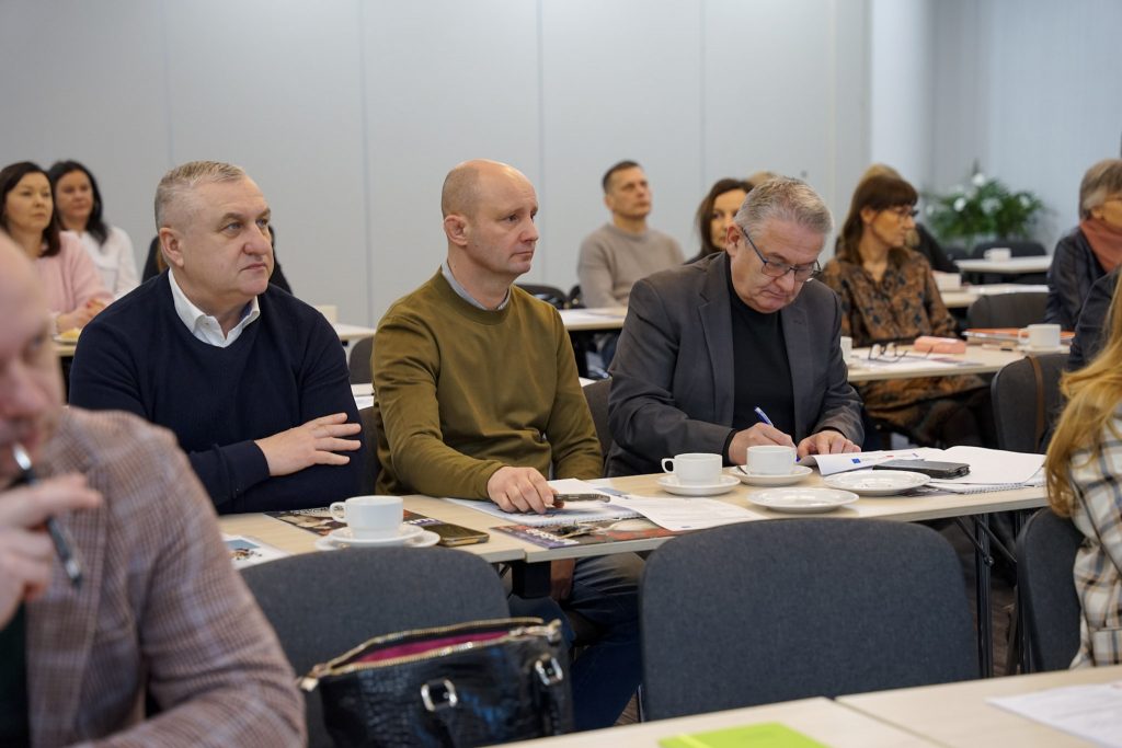 Kilku mężczyzn siedzi przy stole; jeden pisze w notesie, przed nimi filiżanki.