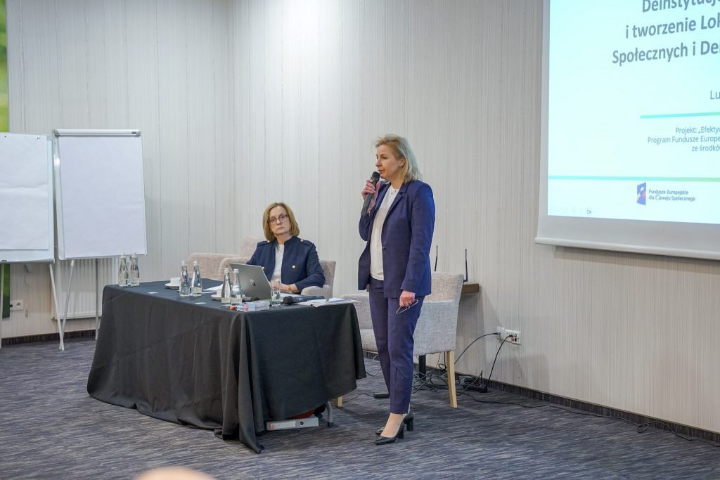 Dwie kobiety. Jedna siedzi, druga stoi, jedna mówi do mikrofonu, w tle slajd.