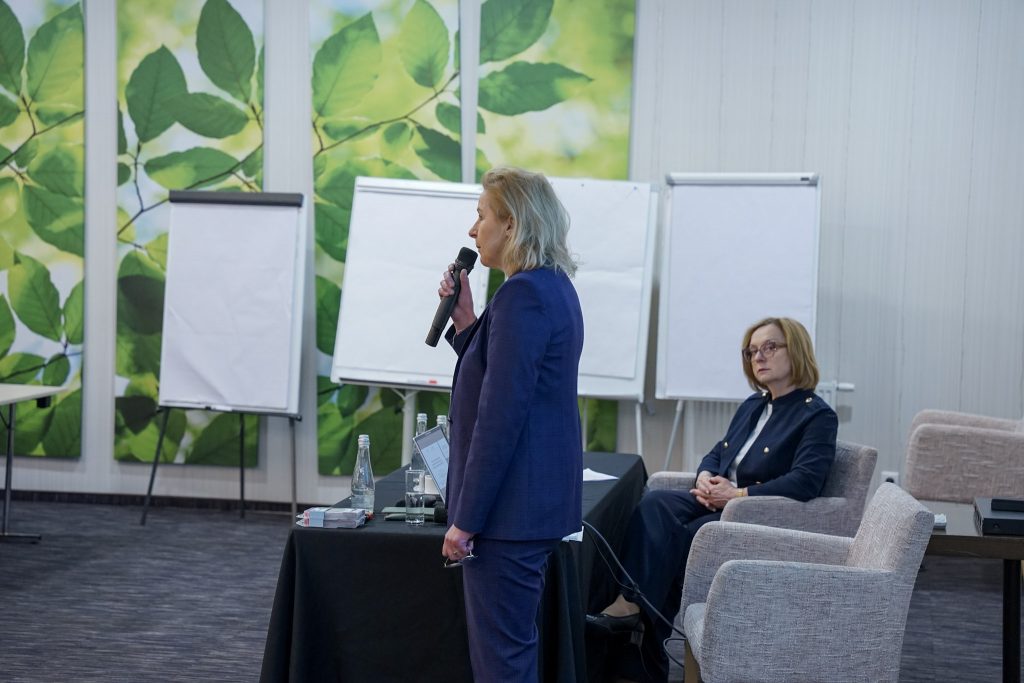 Na pierwszym planie kobieta stojąca bokiem do aparatu przemawia do uczestników, trzymając mikrofon. Ubrana jest w granatowy garnitur, stoi obok stolika z wodą i materiałami szkoleniowymi. W tle, po prawej stronie, druga kobieta siedzi na fotelu, uważnie słuchając wystąpienia. Również ubrana jest w formalny strój. Za nimi znajdują się tablice typu flipchart, a ściany sali ozdobione są grafikami z motywem zielonych liści, co nadaje przestrzeni spokojny, profesjonalny charakter.