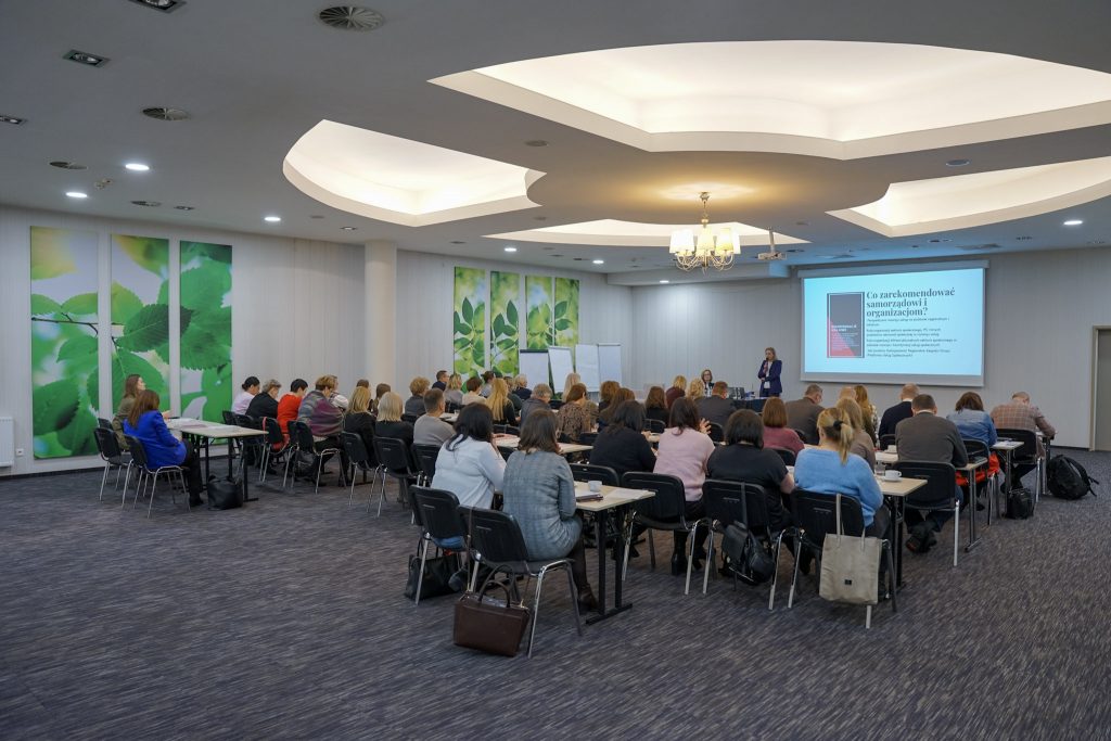 Sala konferencyjna z uczestnikami siedzącymi przy stołach, widok od tyłu.