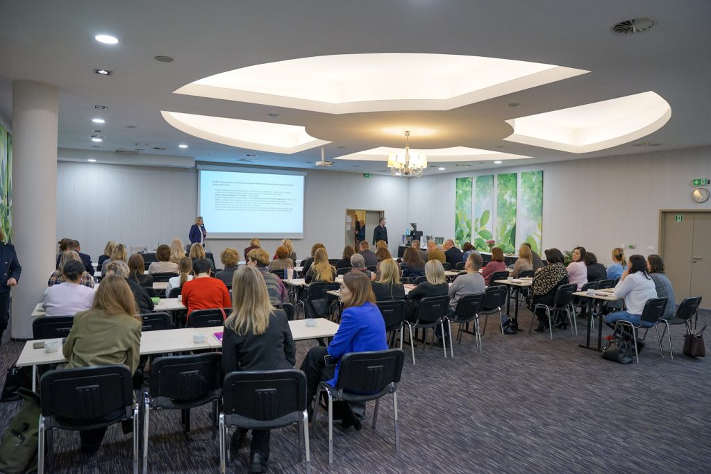 Sala konferencyjna z uczestnikami siedzącymi przy stołach, widok od tyłu.